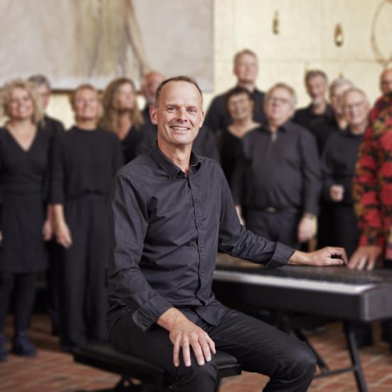 Billedet forestiller Jesper Bauder der sidder ved keybordet med et kor i baggrunden.