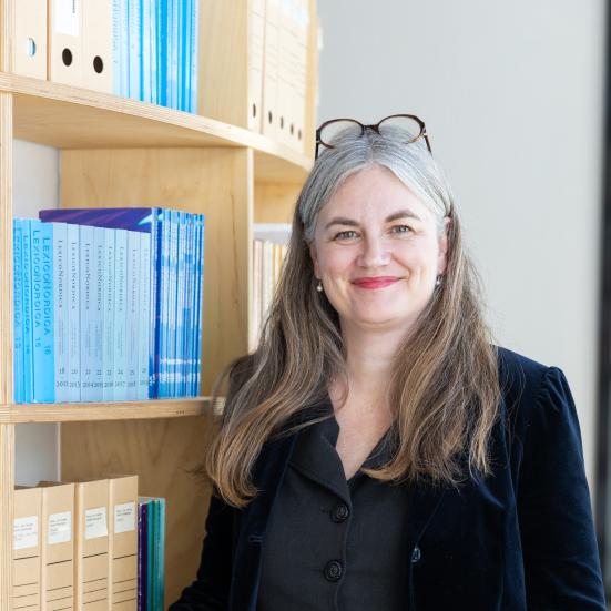 Billedet viser en smilende Marianne Rathje ved siden af en fyldt reol