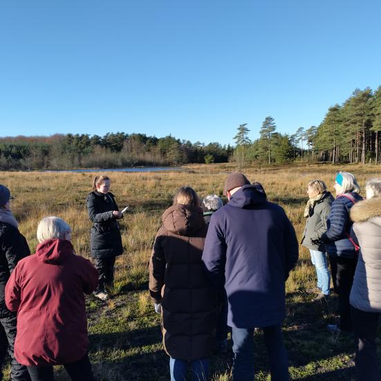 Billedet viser en gruppe mennesker ude i naturen, som står i en halvcirkel omkring en person, der læser op af en bog
