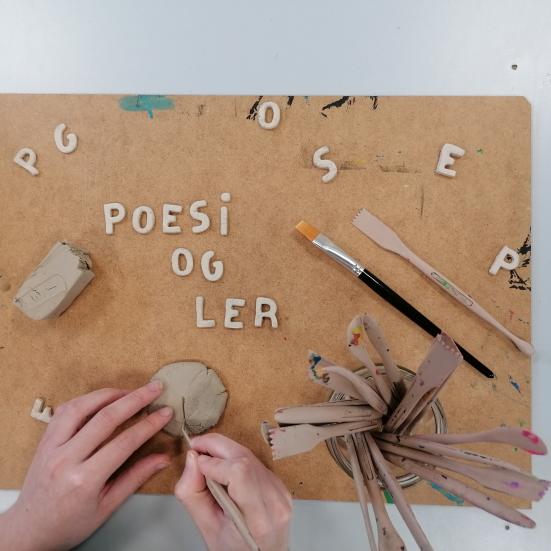 Fotoet viser et par hænder i gang med at forme bogstaver i ler på en masonitplade