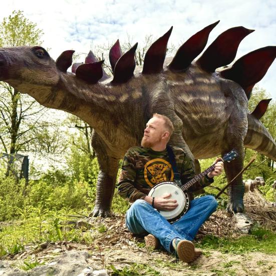 Billedet viser Morten, som siddende spiller banjo foran en dinosaur