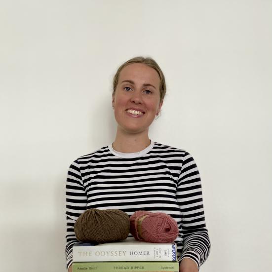 Fotoet viser en smilende Johanne Bisgaard med en stak bøger og et par garnnøgler i favnen