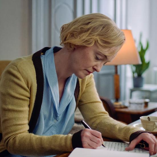 Billedet forestiller en kvinde bøjet over et bord, mens hun skriver på et stykke papir.