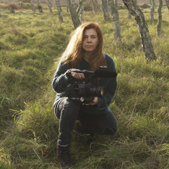 Billedet forestiller en kvinde siddende i græsset mellem unge træer.