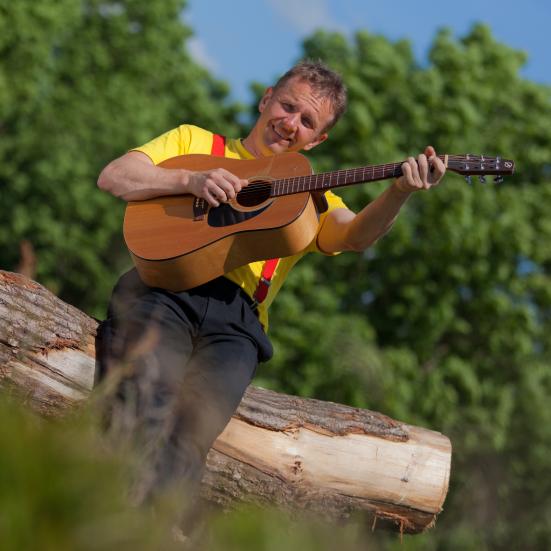 Billedet viser Michel Back siddende på en træstamme med en guitar i hænderne