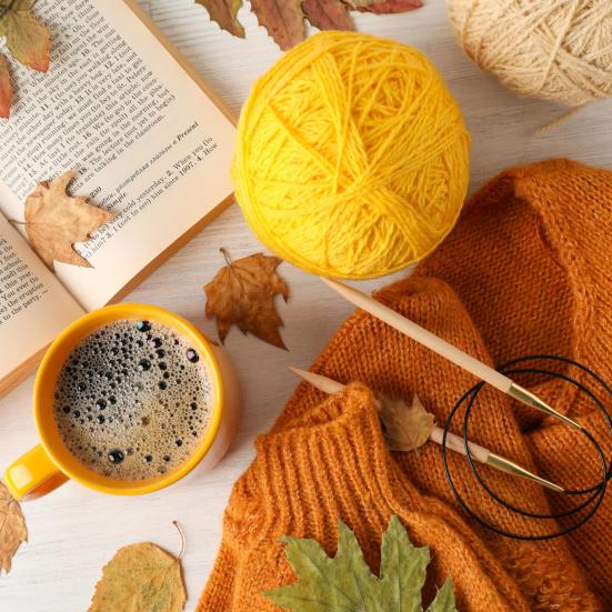 Billedet viser et strikketøj i orange og gule farver sammen med en kop kaffe og en bog på et bord