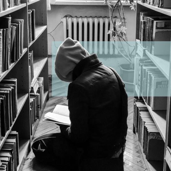 Sort-hvid foto af en person med hætte, der læser på gulvet i et bibliotek.