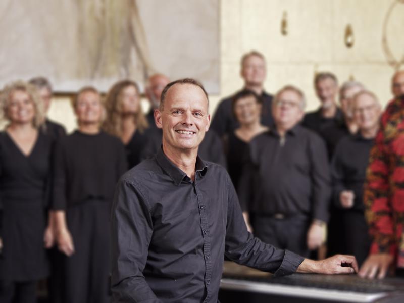 Billedet forestiller Jesper Bauder der sidder ved keybordet med et kor i baggrunden.