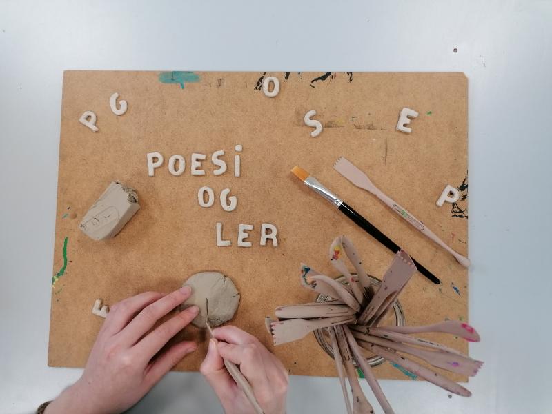 Fotoet viser et par hænder i gang med at forme bogstaver i ler på en masonitplade