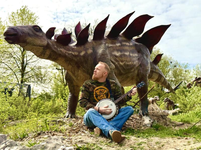 Billedet viser Morten, som siddende spiller banjo foran en dinosaur