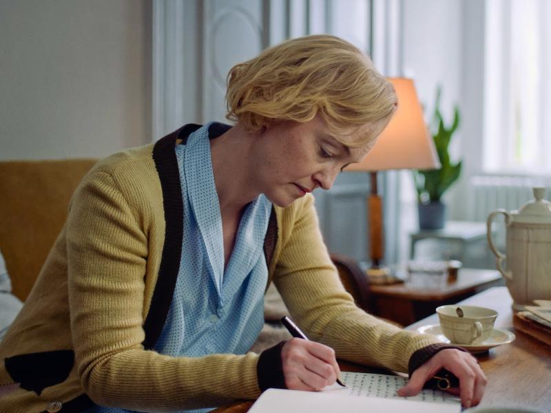 Billedet forestiller en kvinde bøjet over et bord, mens hun skriver på et stykke papir.