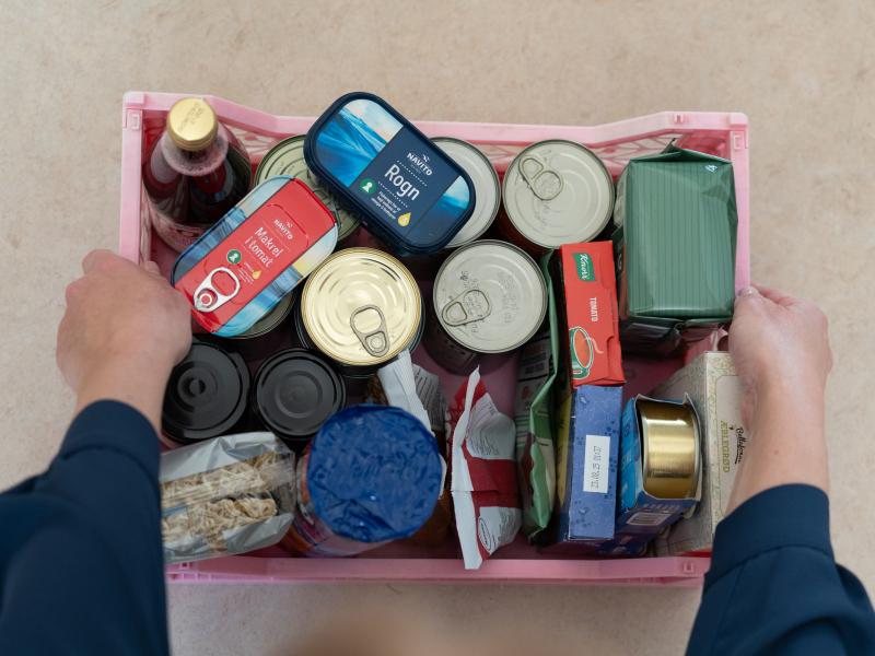 Billedet viser et par hænder, der holder en kasse med dåsemad og andre fornødenheder