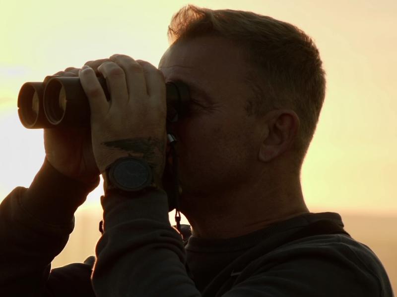 Billedet forestiller en mand der står med en kikkert. Bag ham er solen, og himlen har en gylden farve.