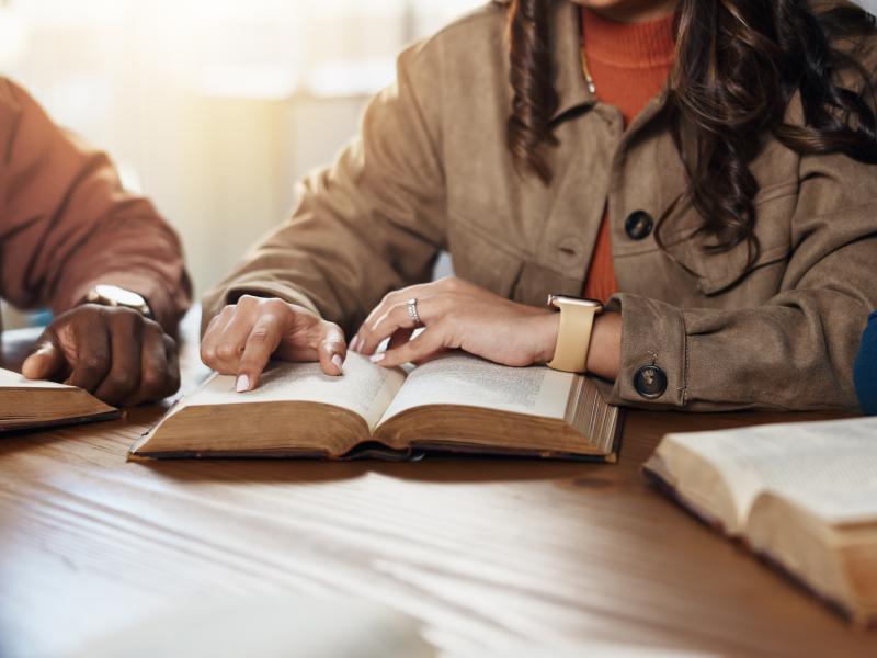 Billedet viser tre opslåede bøger på et bord, samt hænderne på dem, der sidder med bøgerne