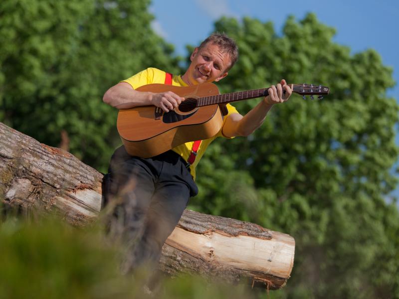 Billedet viser Michel Back siddende på en træstamme med en guitar i hænderne