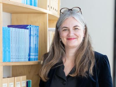 Billedet viser en smilende Marianne Rathje ved siden af en fyldt reol