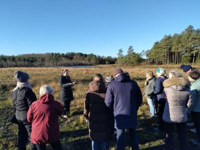 Billedet viser en gruppe mennesker ude i naturen, som står i en halvcirkel omkring en person, der læser op af en bog