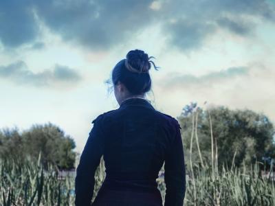 Billedet forestiller en kvinde set fra ryggen. Hun er iført en gammeldags mørk kjole og har sit mørke hår sat op i en knold.