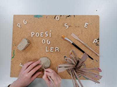 Fotoet viser et par hænder i gang med at forme bogstaver i ler på en masonitplade