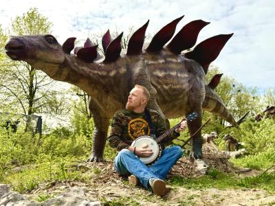 Billedet viser Morten, som siddende spiller banjo foran en dinosaur