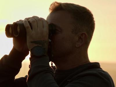 Billedet forestiller en mand der står med en kikkert. Bag ham er solen, og himlen har en gylden farve.