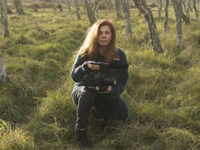 Billedet forestiller en kvinde siddende i græsset mellem unge træer.