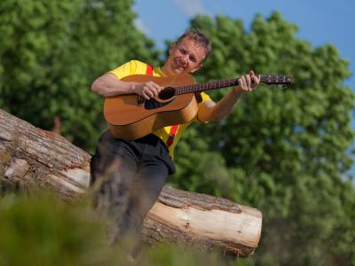 Billedet viser Michel Back siddende på en træstamme med en guitar i hænderne