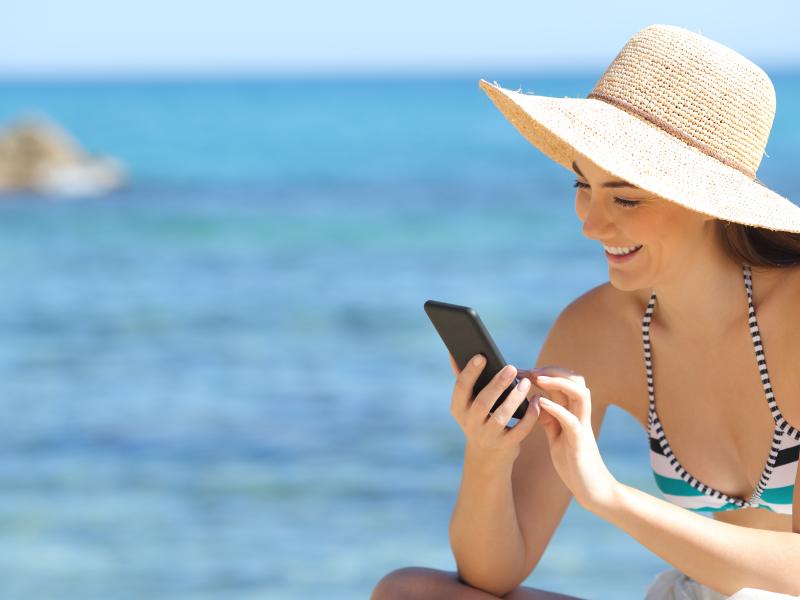 Billedet forestiller en smilende kvinde iført bikini og hat på stranden. Hun smiler, mens hun kigger på sin telefon. I baggrunden kan man se havet.