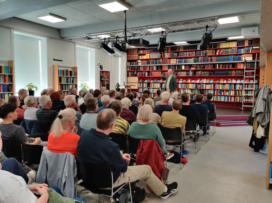 Billedet viser Salonen på Varde Bibliotek under et arrangement. Det er taget ud over stolerækkerne, som er fyldt med voksne mennesker