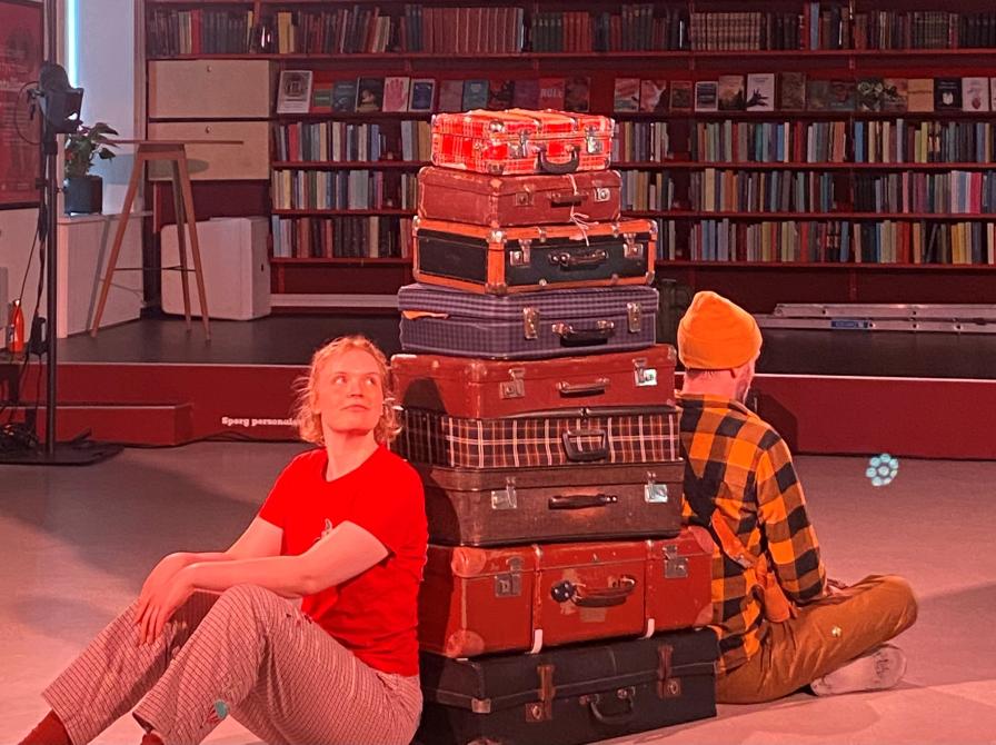 Billedet viser to dansere som sidder på hver sin side af en stor stak kufferter på Varde Bibliotek