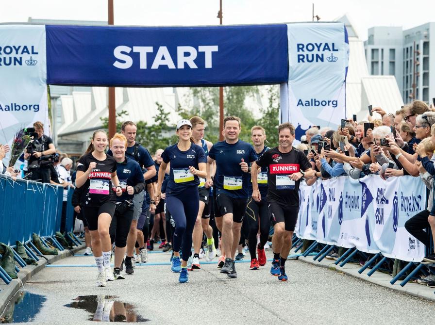 Løbere sammen med Dronning Mary passerer startporten til Royal Run i Aalborg, mens tilskuere hepper fra siderne.