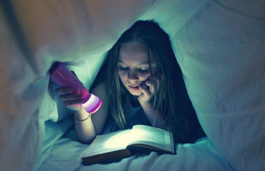 Billedet forestiller en pige der har lavet en hule med sin dyne. Hun ligger og læser i en bog som hun lyser op med en lommelygte.