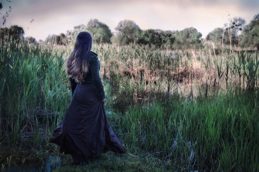 Billedet forestiller en ung kvinde set fra ryggen. Hun er iført en lang, mørk kjole og står og ser ud over en sø.