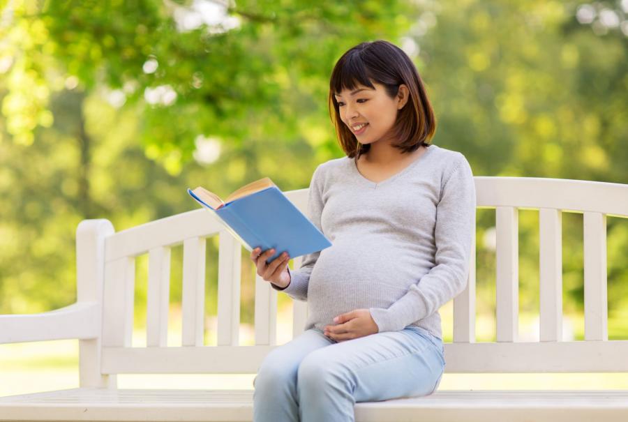 Gravid kvinde sidder smilende på en hvid bænk. Hun læser i en bog. I baggrunden er der træer.