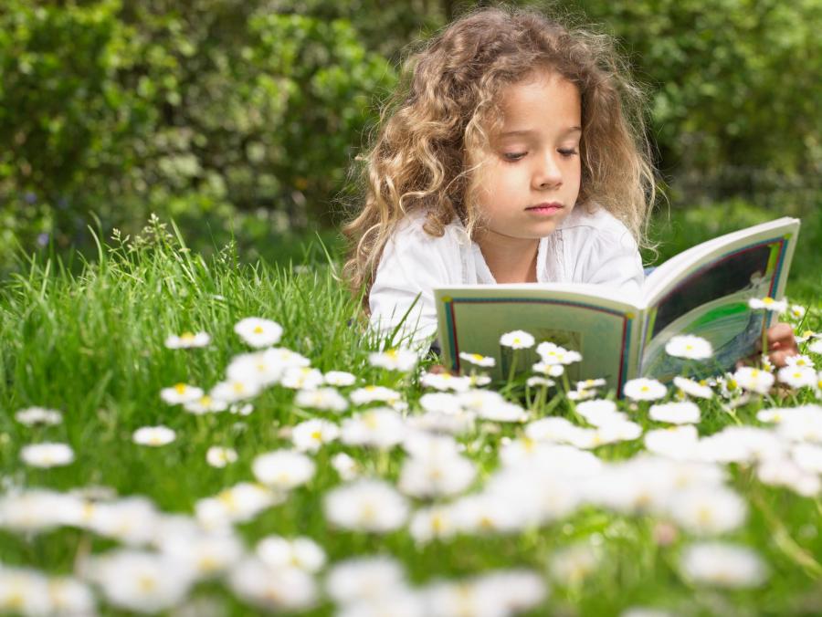 Billedet forestiller en lille pige med lyst, krøllet hår. Hun ligger på maven på en græsplæne med hvide blomster. Hun holder en bog op foran sig og læser.