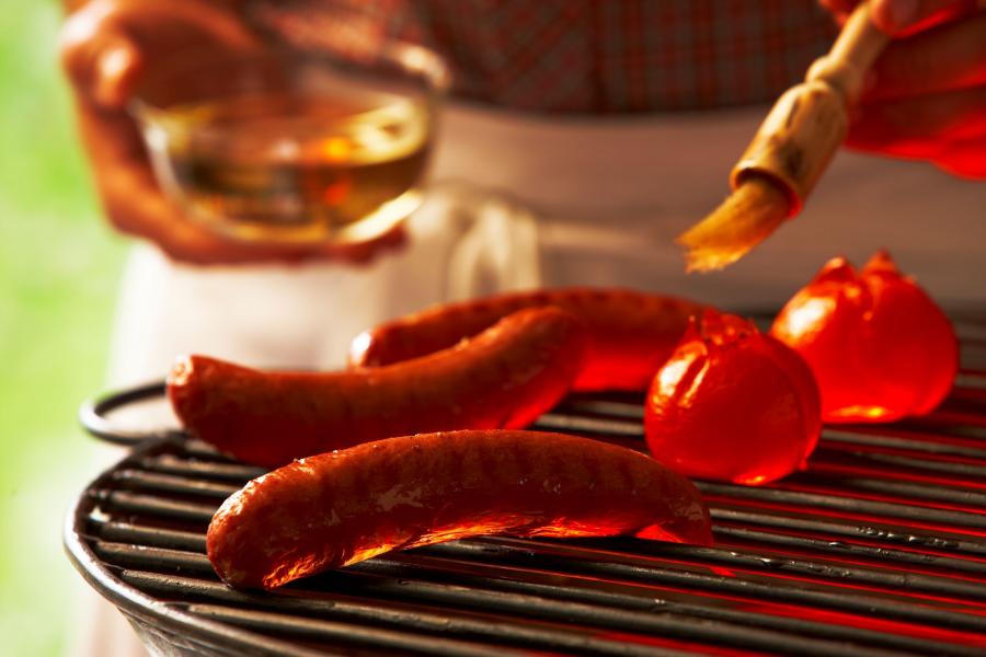 Billedet forestiller et nærbillede af en grill. Der ligger tre røde pølser på grillen samt to tomater. Man kan se et par hænder, der er i gang med at pensle maden.