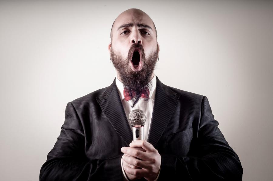 Billedet forestiller en mand med skæg. Han synger dybfølt ind i en mikrofon, som han holder med begge hænder. 