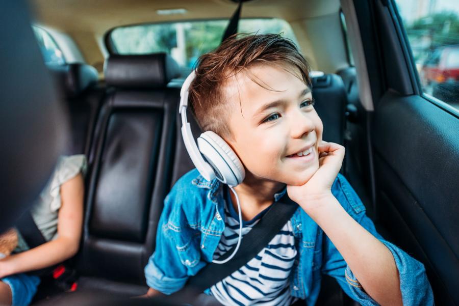 Billedet forestiller en dreng, der sidder på bagsædet af en bil. Han kigger smilende ud af vinduet, mens han hviler hagen i den ene hånd.  Han har hvide hørertelefoner på.