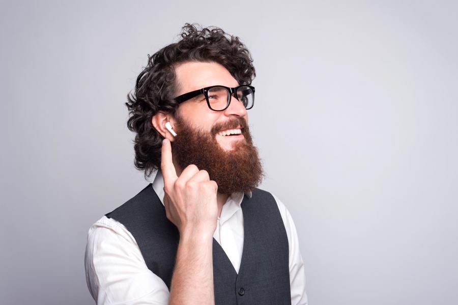 Billedet forestiller en ung mand med et stort, busket skæg. Han har earplugs i ørerne og lytter med et stort smil.