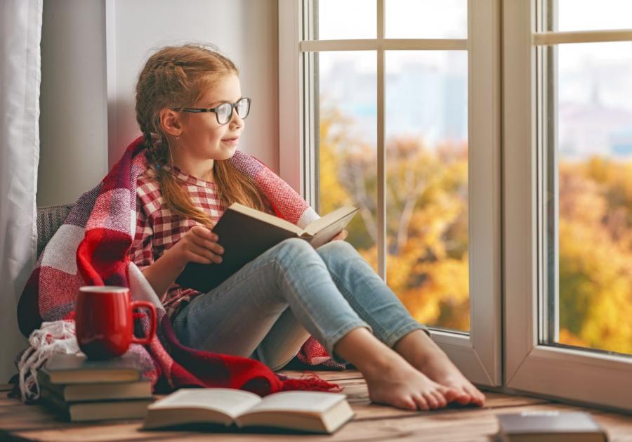 Billedet forestiller en pige på cirka 8 år der sidder i et vindue med en bog. Hun har et tæppe over skuldrene og et rødt krus stående ved sin side.